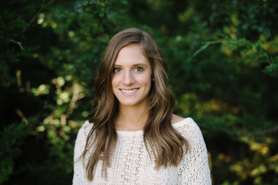 A young woman with long brown hair wearing a white knitted sweater stands outdoors in front of green foliage, smiling at the camera. by Justin Salem Meyer