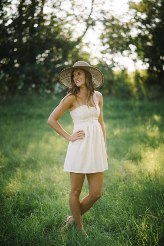 A woman wearing a sunhat and a white sleeveless dress stands smiling in a sunlit grassy field, with trees and greenery in the background. by Justin Salem Meyer