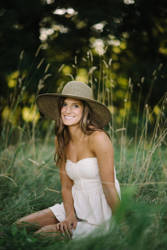 A young woman in a white strapless dress and wide-brimmed straw hat sits in tall grass, smiling, with a blurred background of greenery and sunlight filtering through trees. by Justin Salem Meyer
