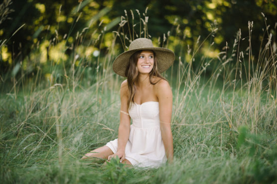 A woman in a white strapless dress and wide-brimmed hat sits in tall grass, smiling, with a blurred background of green trees and sunlight filtering through. by Justin Salem Meyer