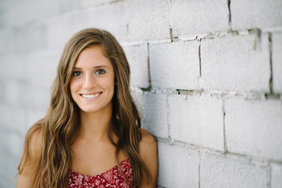 A young woman with long brown hair and a red patterned top smiles while leaning against a white brick wall. by Justin Salem Meyer