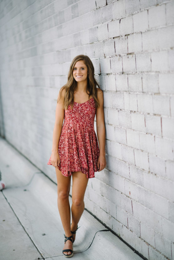 A young woman in a red floral dress and black sandals stands, smiling, against a light gray brick wall on a sidewalk. Her long brown hair is down, and she has a relaxed, casual pose. by Justin Salem Meyer