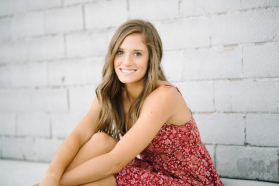 A young woman with long brown hair wearing a sleeveless red floral dress sits against a white brick wall, smiling at the camera. by Justin Salem Meyer