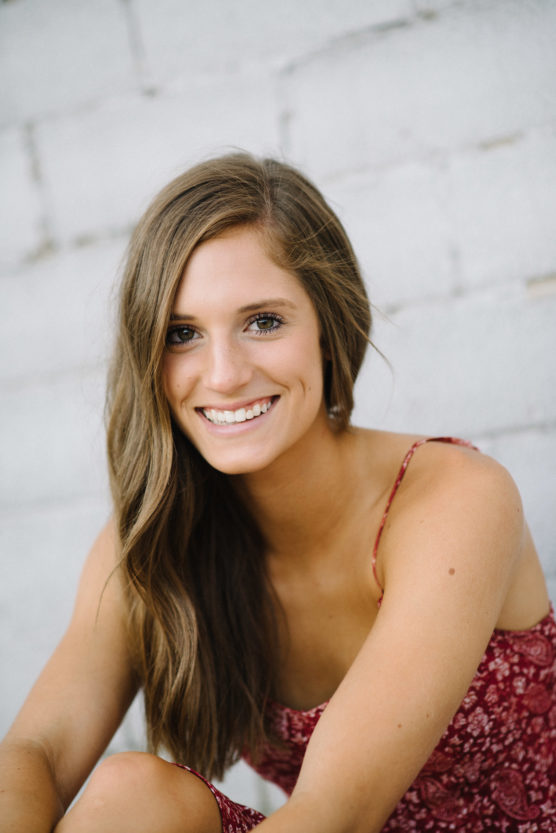 A young woman with long brown hair, wearing a sleeveless red patterned dress, sits and smiles at the camera in front of a light-colored brick wall. by Justin Salem Meyer