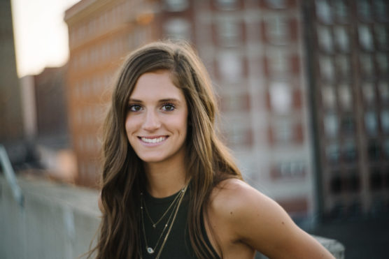 A young woman with long brown hair and a sleeveless dark top smiles at the camera, standing outdoors with blurred city buildings in the background. by Justin Salem Meyer