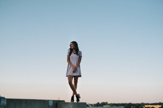 A young woman in a short, light-colored dress and black shoes stands on a concrete ledge outdoors, smiling with her arms loosely crossed. The sky is clear and blue, and trees are visible in the distance. by Justin Salem Meyer