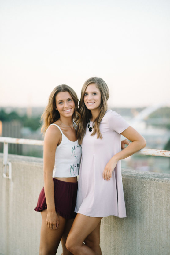 Two young women stand side by side outdoors, smiling at the camera. One wears a white tank top and maroon shorts, and the other wears a light pink dress. They lean against a concrete barrier with a cityscape in the background. by Justin Salem Meyer