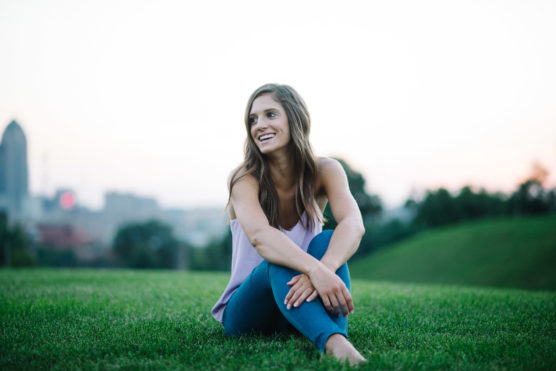 A young woman with long brown hair sits cross-legged on grass in a park, smiling and looking slightly to the side. The background shows a blurred city skyline at dusk. by Justin Salem Meyer