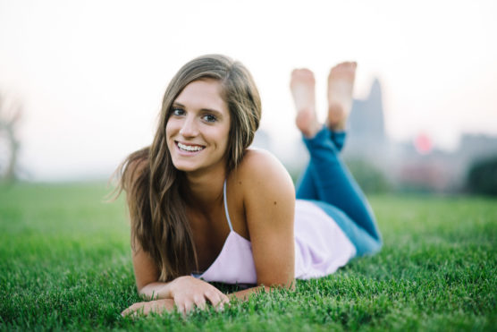 A young woman with long brown hair and a light purple top lies on her stomach in the grass, smiling at the camera with her feet up and crossed, with a blurred cityscape in the background. by Justin Salem Meyer