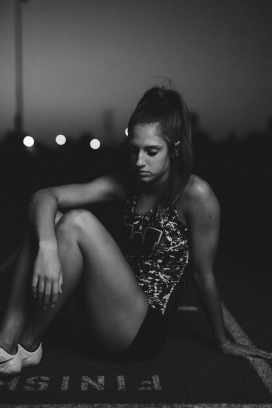 A young woman sits on a track at dusk, looking down thoughtfully with her knees bent. She wears athletic clothes and appears to be resting near the FINISH line. The image is in black and white. by Justin Salem Meyer