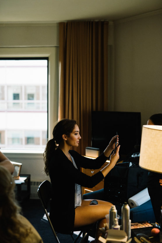 A woman sits on a chair in a dimly lit room, holding up a phone or mirror while looking at it. A large window and beige curtains are in the background, with lamps and beauty products on a nearby table. by Justin Salem Meyer
