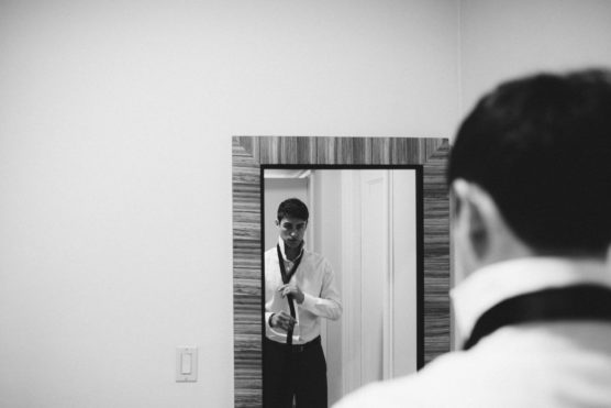 A man stands in front of a mirror, adjusting his tie while getting dressed. The image is in black and white, and the scene is captured from behind and slightly to the side of the man. by Justin Salem Meyer