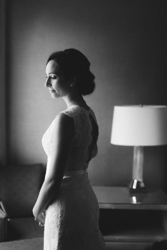 A woman in a lace wedding dress stands in profile, eyes closed, in a softly lit room with a table and lamp in the background. The photo is in black and white. by Justin Salem Meyer