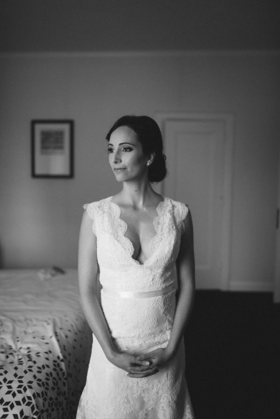 A woman in a white lace wedding dress stands indoors, hands clasped, gazing thoughtfully to the side. The room is softly lit, with a bed and framed picture in the background. The image is in black and white. by Justin Salem Meyer