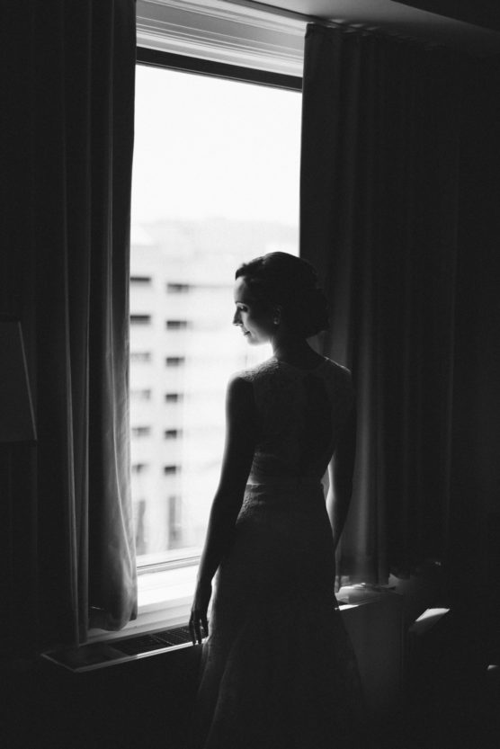 A woman in a sleeveless dress stands by a window, looking outside. The scene is in black and white, with soft light illuminating her profile and creating a serene, contemplative mood. by Justin Salem Meyer