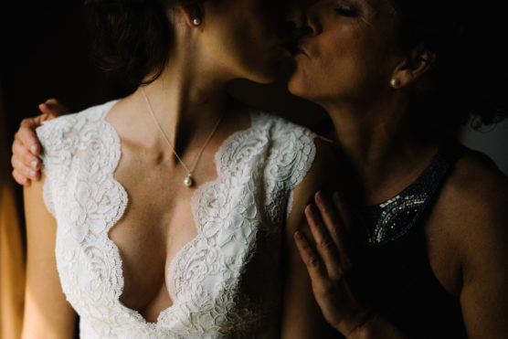 Two women share an intimate kiss. One wears a white lace dress with a pearl necklace, while the others hand rests gently on her shoulder, both partly in shadow. by Justin Salem Meyer