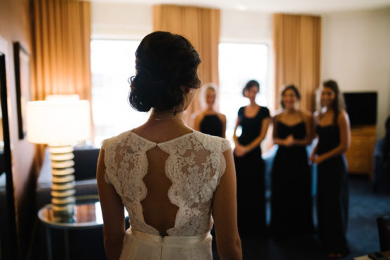 A bride in a lace wedding dress stands with her back to the camera, facing four bridesmaids in dark dresses who are smiling in a softly lit room with large curtained windows. by Justin Salem Meyer