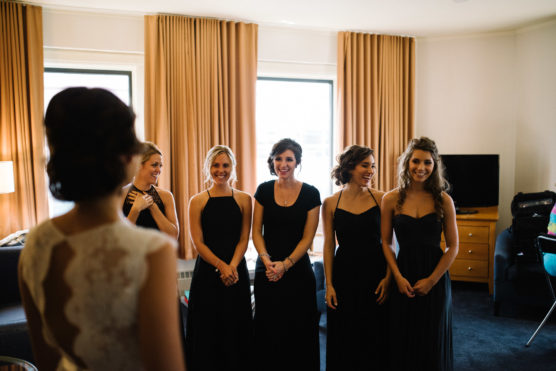 Five women in black dresses smile and face a bride in a white lace gown inside a well-lit room with tan curtains and modern furniture. The atmosphere is joyful and celebratory. by Justin Salem Meyer