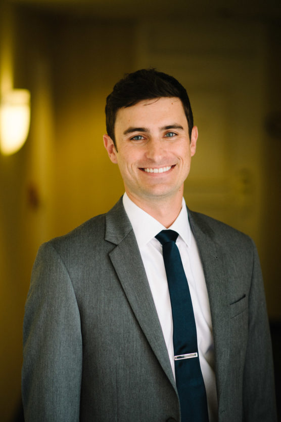 A young man with short dark hair, wearing a gray suit, white shirt, and dark tie, smiles at the camera in a softly lit indoor setting. by Justin Salem Meyer