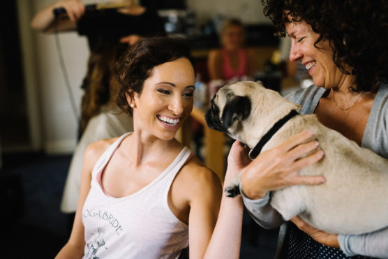 A woman in a tank top smiles at a small pug dog being held by another woman, both looking happy indoors. Others are blurred in the background, creating a cheerful, casual atmosphere. by Justin Salem Meyer