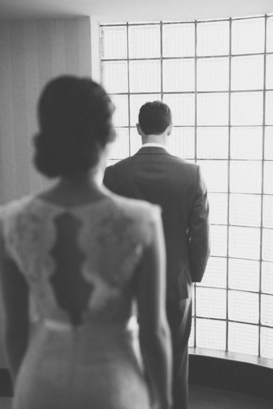 A black and white photo of a woman in a lace dress standing behind a man in a suit, both facing a wall of glass bricks. The mans back is to the camera, and the image is softly focused. by Justin Salem Meyer