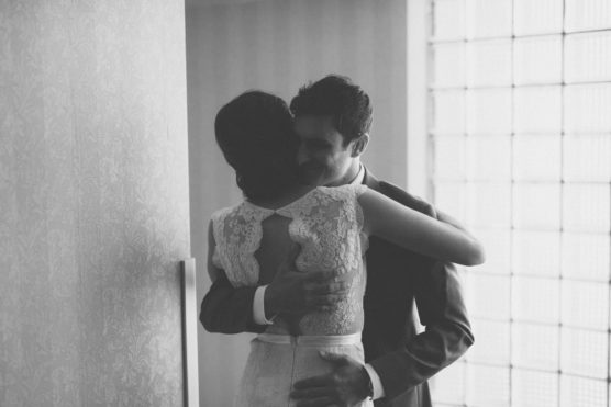 A black and white photo of a couple embracing closely indoors; the woman wears a lace dress and the man wears a suit. Their faces are partly hidden in the hug, and soft light filters in through a window. by Justin Salem Meyer
