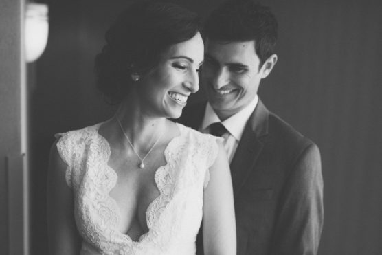 A black-and-white photo of a couple on their wedding day. The woman in a lace dress smiles and looks down, while the man in a suit stands behind her, smiling warmly. by Justin Salem Meyer