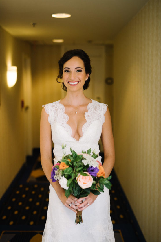 A smiling bride in a white lace dress holds a colorful bouquet of flowers, standing in a warmly lit hallway with patterned carpet and cream-colored walls. by Justin Salem Meyer