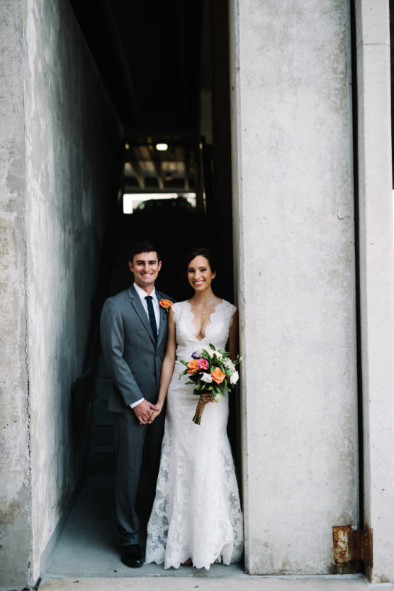 A bride in a white lace gown holding a bouquet stands next to a groom in a gray suit with a peach boutonniere. They are smiling, standing in a concrete doorway with stairs and shadows behind them. by Justin Salem Meyer