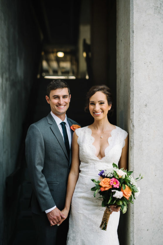 A smiling bride in a white lace dress holds a colorful bouquet while standing beside a groom in a gray suit with an orange boutonniere. They stand close together in a concrete corridor, holding hands. by Justin Salem Meyer