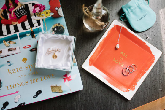 A flat lay of a Kate Spade book, a jewelry box with a gold pendant, a red dish with rings and a necklace, a glass bottle, and a blue Tiffany & Co. pouch on a dark table. by Justin Salem Meyer
