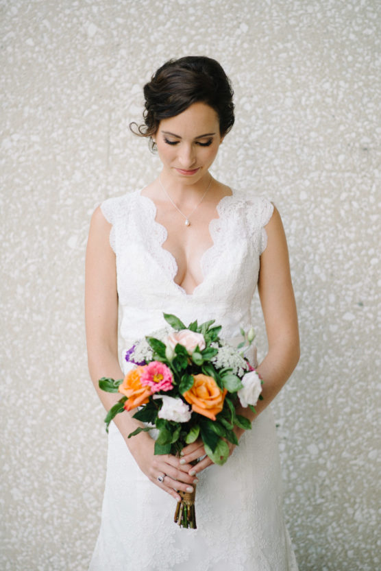 A woman in a white lace wedding dress holds a colorful bouquet of flowers and looks down, standing against a light textured background. by Justin Salem Meyer