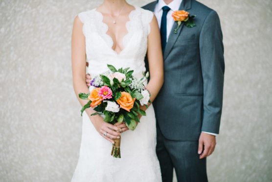 A bride in a white dress holding a colorful bouquet stands next to a groom in a gray suit and blue tie with an orange boutonnière, both facing forward against a neutral background. by Justin Salem Meyer