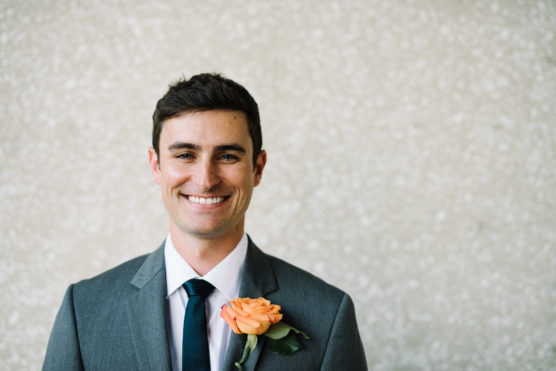 A man in a gray suit and dark tie smiles at the camera, wearing a peach-colored rose boutonniere on his jacket, standing in front of a light textured background. by Justin Salem Meyer