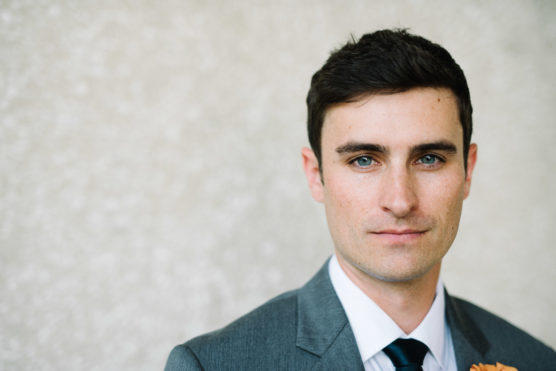 A young man with short dark hair and blue eyes, wearing a gray suit, white shirt, and dark tie, stands in front of a light, textured background, looking directly at the camera with a serious expression. by Justin Salem Meyer