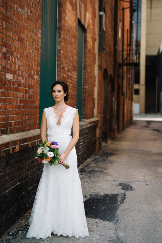 A bride in a white lace gown holding a colorful bouquet stands in a brick alleyway, looking slightly to the side with a calm expression. by Justin Salem Meyer