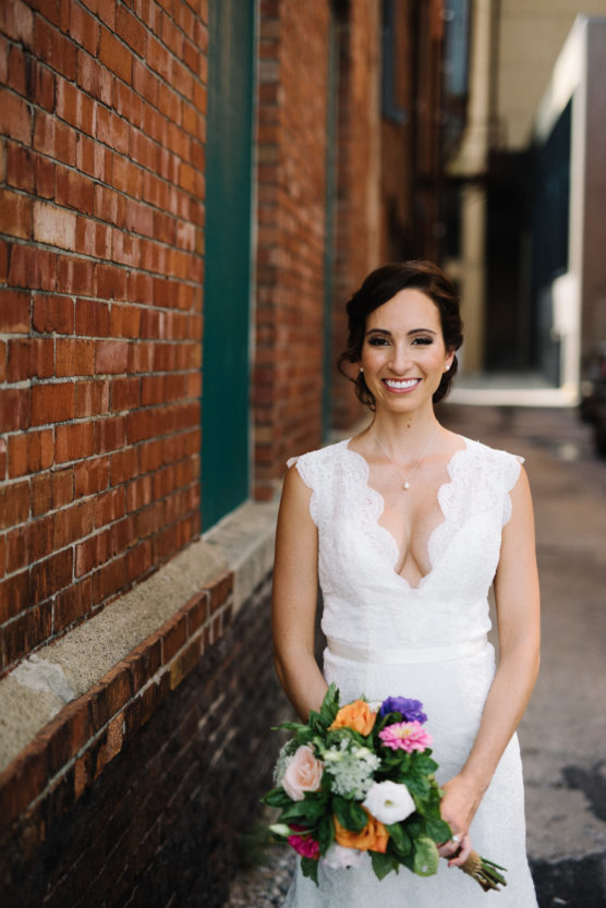 A bride in a white lace dress holds a colorful bouquet and smiles, standing by a brick wall in an urban alleyway. by Justin Salem Meyer