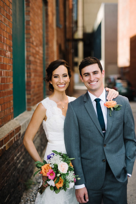 A smiling bride in a white lace dress holds a colorful bouquet and stands with her arm around a groom in a gray suit and navy tie. They pose together in a brick alleyway on a sunny day. by Justin Salem Meyer