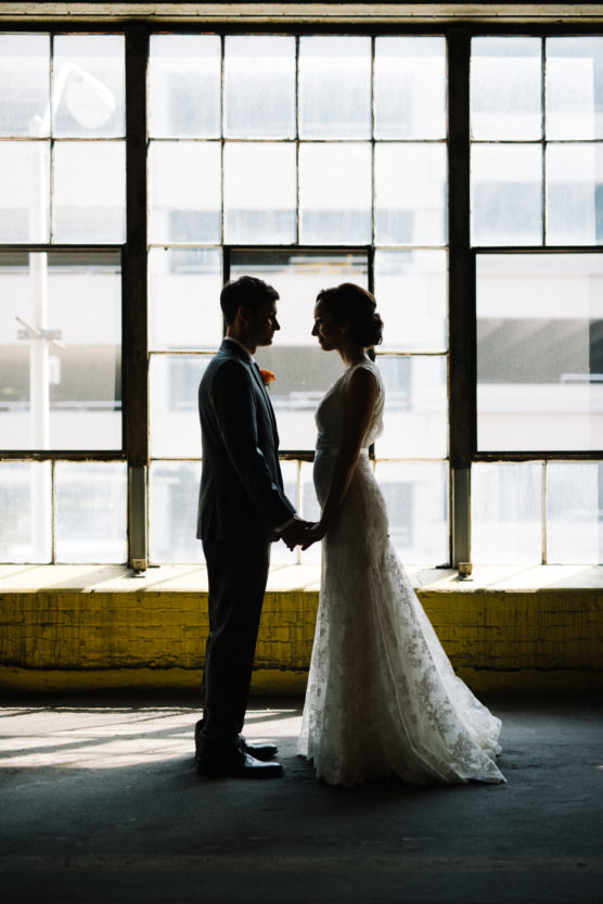 A bride and groom stand facing each other, holding hands, silhouetted against large industrial windows with daylight streaming in behind them. by Justin Salem Meyer