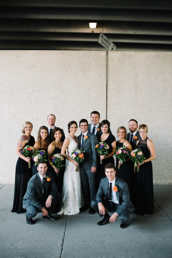 A wedding party poses together. The bride in a white dress and groom in a gray suit stand at the center, surrounded by bridesmaids in black dresses and groomsmen in gray suits, all holding bouquets and smiling. by Justin Salem Meyer