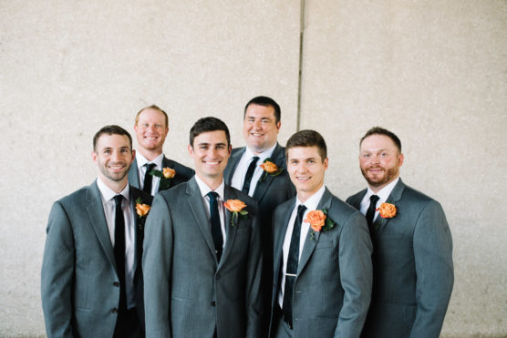 Six men in matching grey suits, white shirts, and dark ties stand smiling together. Three of them wear orange flower boutonnieres. They pose against a neutral, textured wall background. by Justin Salem Meyer