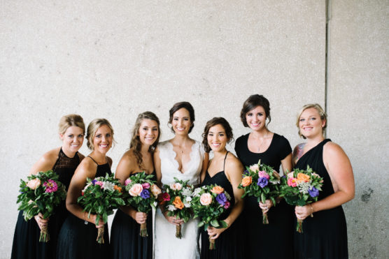 Seven women stand in a row, smiling at the camera. The woman in the center wears a white dress, while the others wear black dresses and hold colorful bouquets of flowers. The background is a plain, textured wall. by Justin Salem Meyer