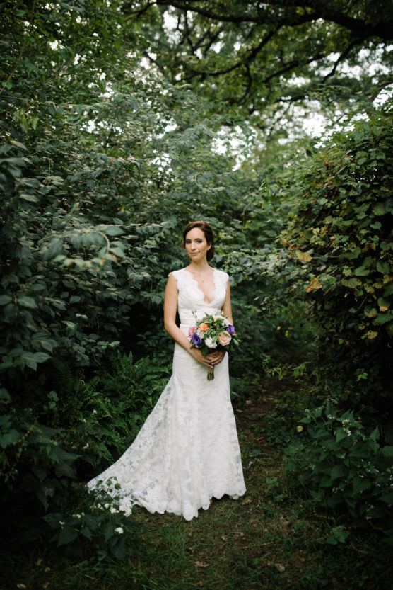 A bride in a white lace gown stands in a lush, green forest holding a colorful bouquet of flowers, surrounded by dense foliage and trees. by Justin Salem Meyer