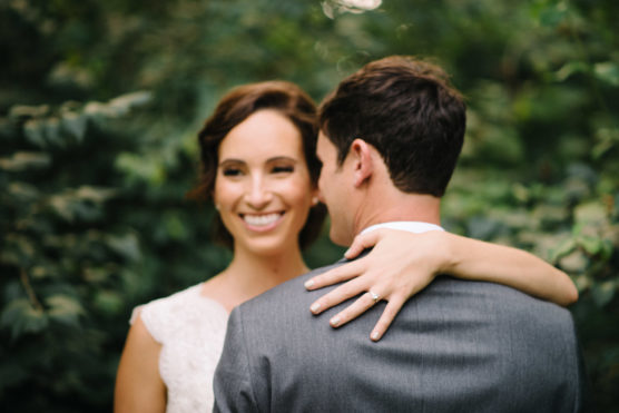 A smiling bride with her arm around a groom’s shoulder, showing her engagement ring, stands outdoors in front of green foliage. The groom is turned away from the camera, while the bride faces forward. by Justin Salem Meyer