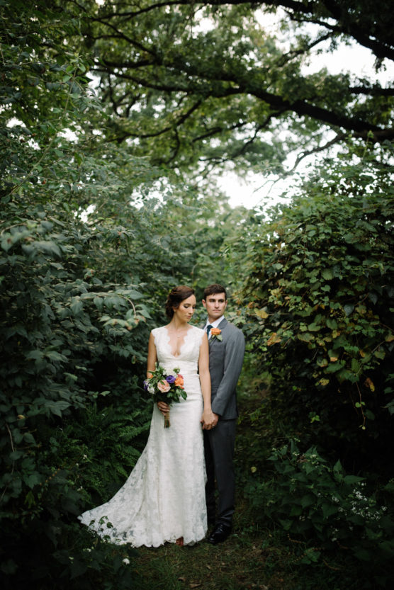 A bride in a white lace gown holding a bouquet stands beside a groom in a gray suit. They hold hands and stand on a green, leafy path surrounded by dense foliage and trees. by Justin Salem Meyer