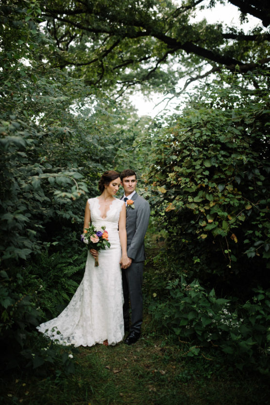 A bride in a white lace dress holding a colorful bouquet stands with a groom in a gray suit in a lush, green, wooded area, surrounded by dense foliage and trees. by Justin Salem Meyer