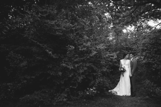 A bride in a long white gown holding a bouquet stands close to a groom in a suit, surrounded by dense, leafy greenery in an outdoor setting. The photo is in black and white. by Justin Salem Meyer