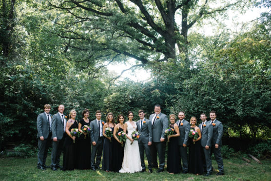 A wedding party poses outdoors in front of lush greenery. The bride and groom stand in the center, surrounded by bridesmaids in black dresses holding bouquets and groomsmen in gray suits. by Justin Salem Meyer
