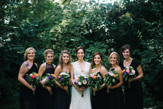 A bride in a white dress stands outdoors with six bridesmaids in black dresses, all holding colorful bouquets, smiling in front of lush green foliage. by Justin Salem Meyer