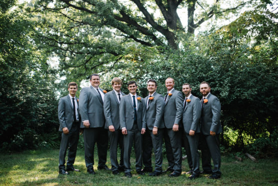 Eight men in matching gray suits with black ties and orange boutonnieres stand in a row outdoors, smiling, with lush green trees and bushes in the background. It appears to be a wedding or formal event. by Justin Salem Meyer